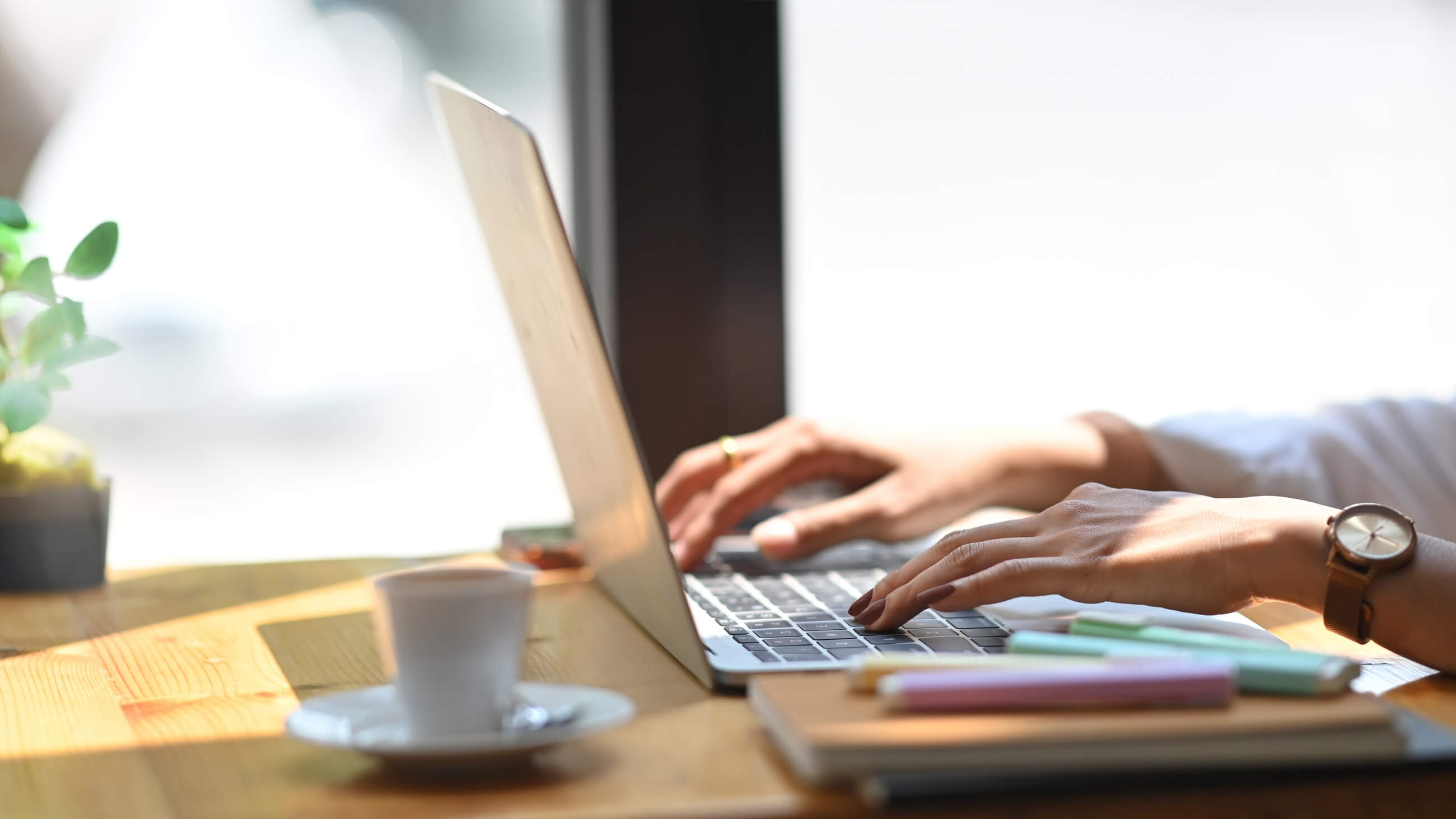 laptop and a coffee mug on a wooden table with hands hovering over the laptop keyboard