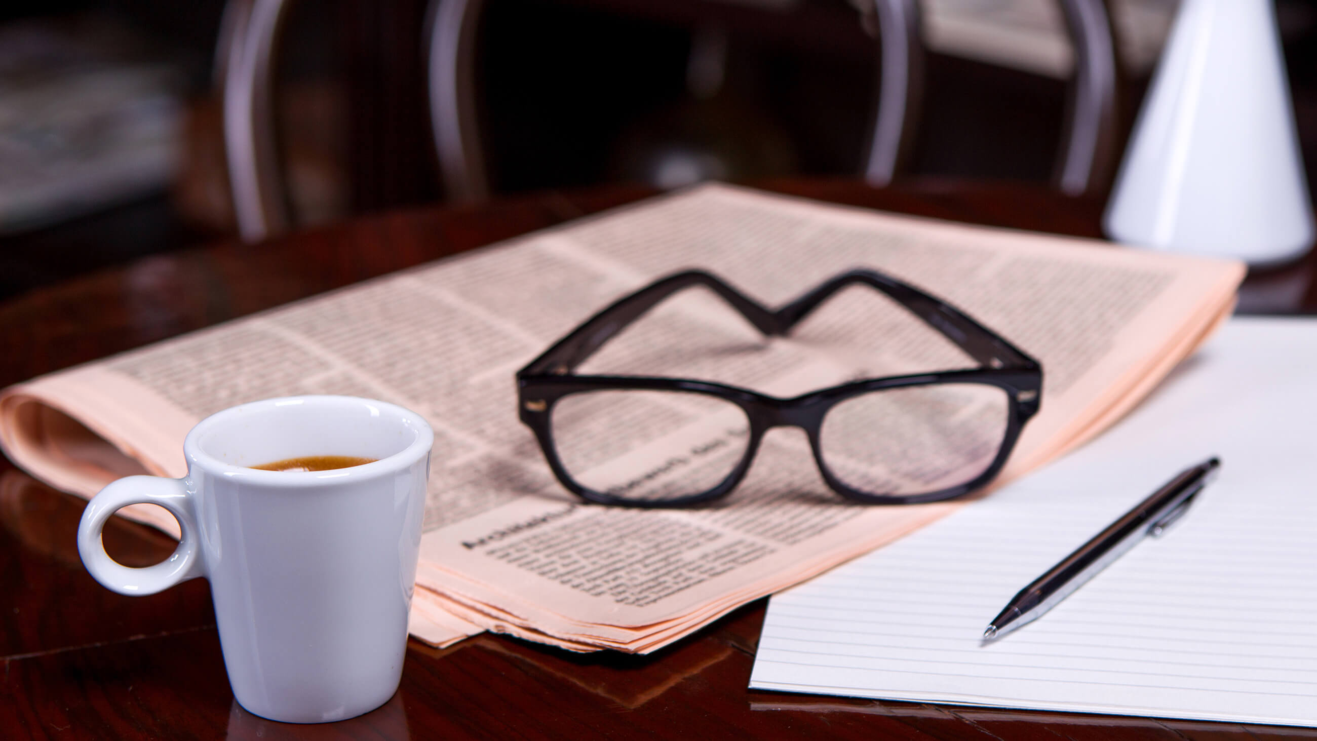 reading glasses over a newspaper with a cup of coffee, paper, and pen next to it